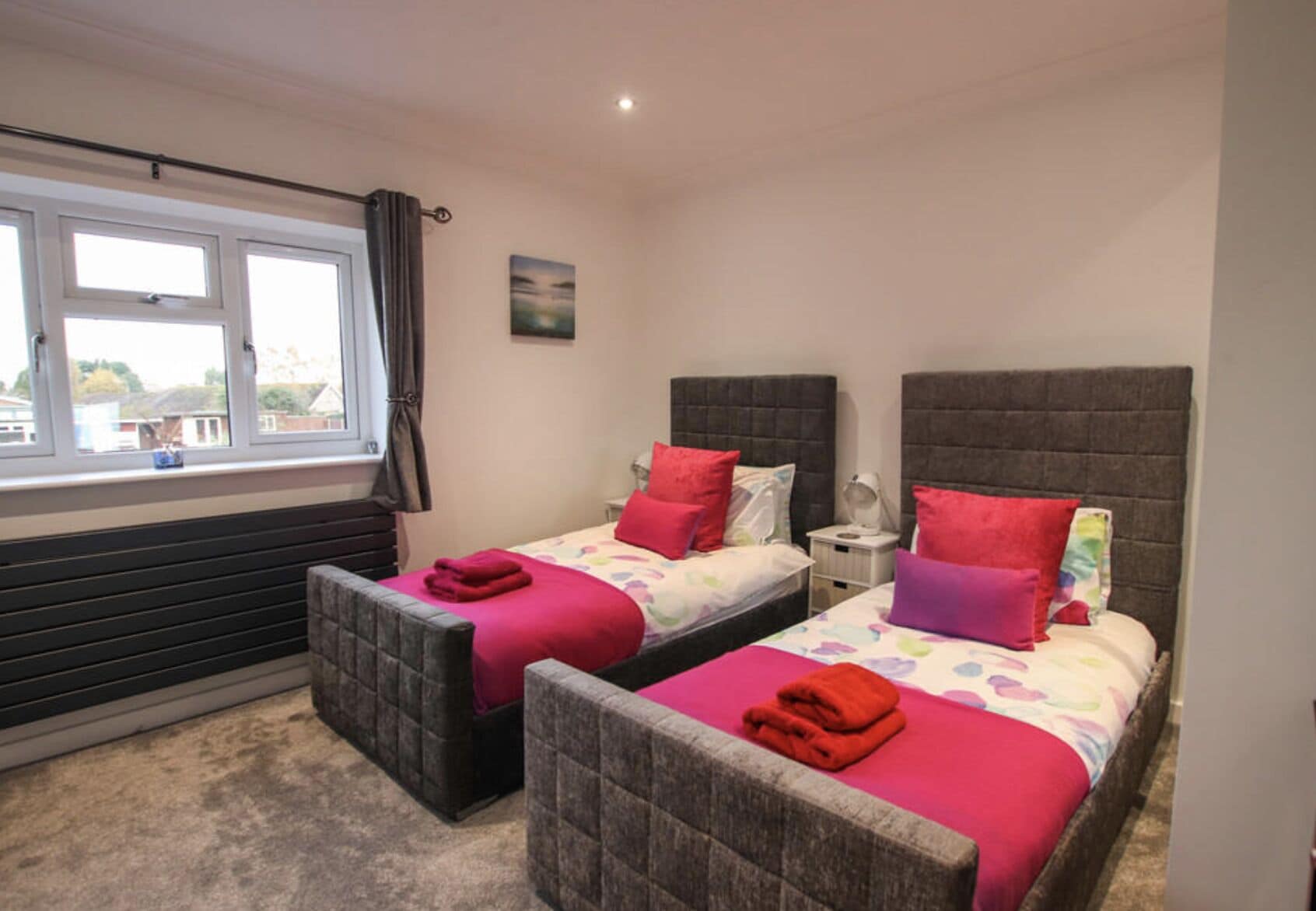A bedroom with two beds, each with a headboard, a window with curtains, and a small bedside table with a lamp.