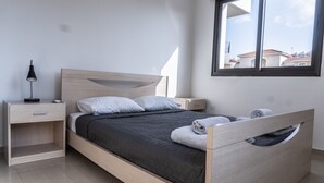A modern bedroom with a wooden bed, a nightstand, a lamp, and a window with a view of buildings. - Luxurious Unique Apartment (Paralimni)