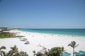 A beach with white sand, clear blue water, and a few palm trees. There are beach umbrellas and lounge chairs.