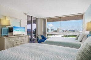 A hotel room with two beds, a TV, a desk with a lamp, and a large window with a city view. - 7th-Floor Oceanfront Condo on Private Beach (Siesta Key)