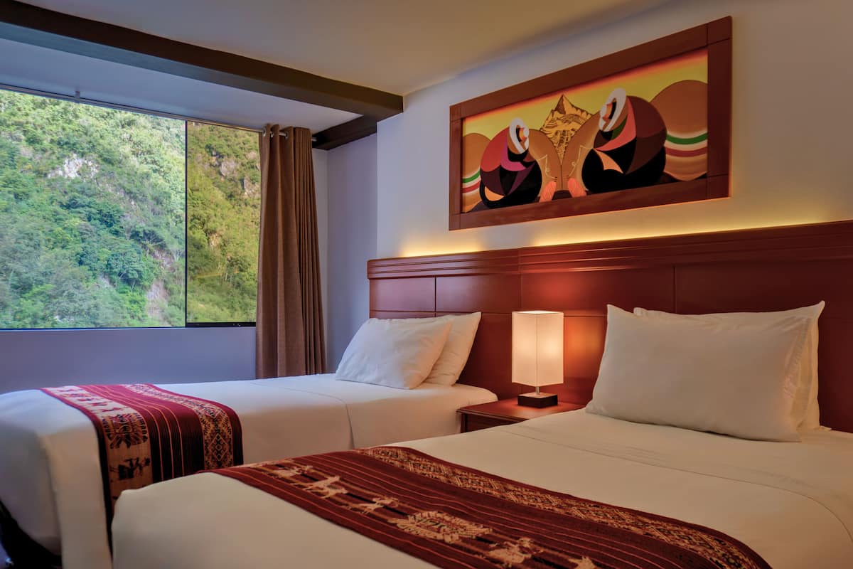 A hotel room with two beds, a large window with a view of greenery, a painting on the wall, and a patterned throw on the beds.