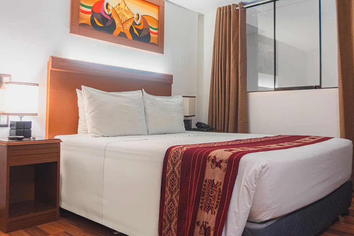 A hotel room with a large bed, a wooden headboard, a nightstand with a lamp, a telephone, and a window with curtains.