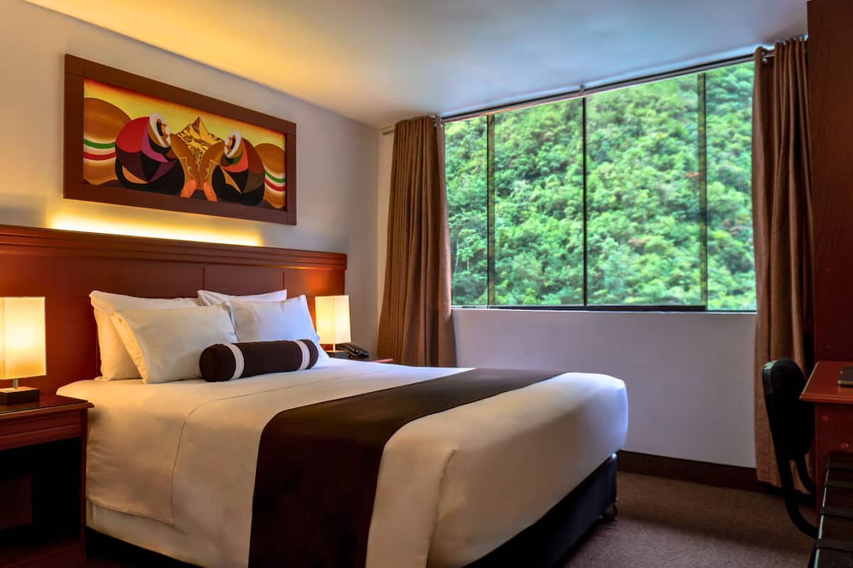 A hotel room with a large bed, two bedside tables with lamps, a painting above the bed, and a window with curtains.