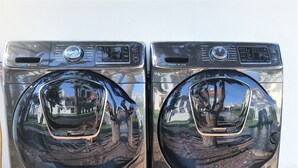 Deux machines à laver à chargement frontal, dotées de panneaux de commande et d’écrans numériques, sont installées à l’extérieur sur une surface pavée.