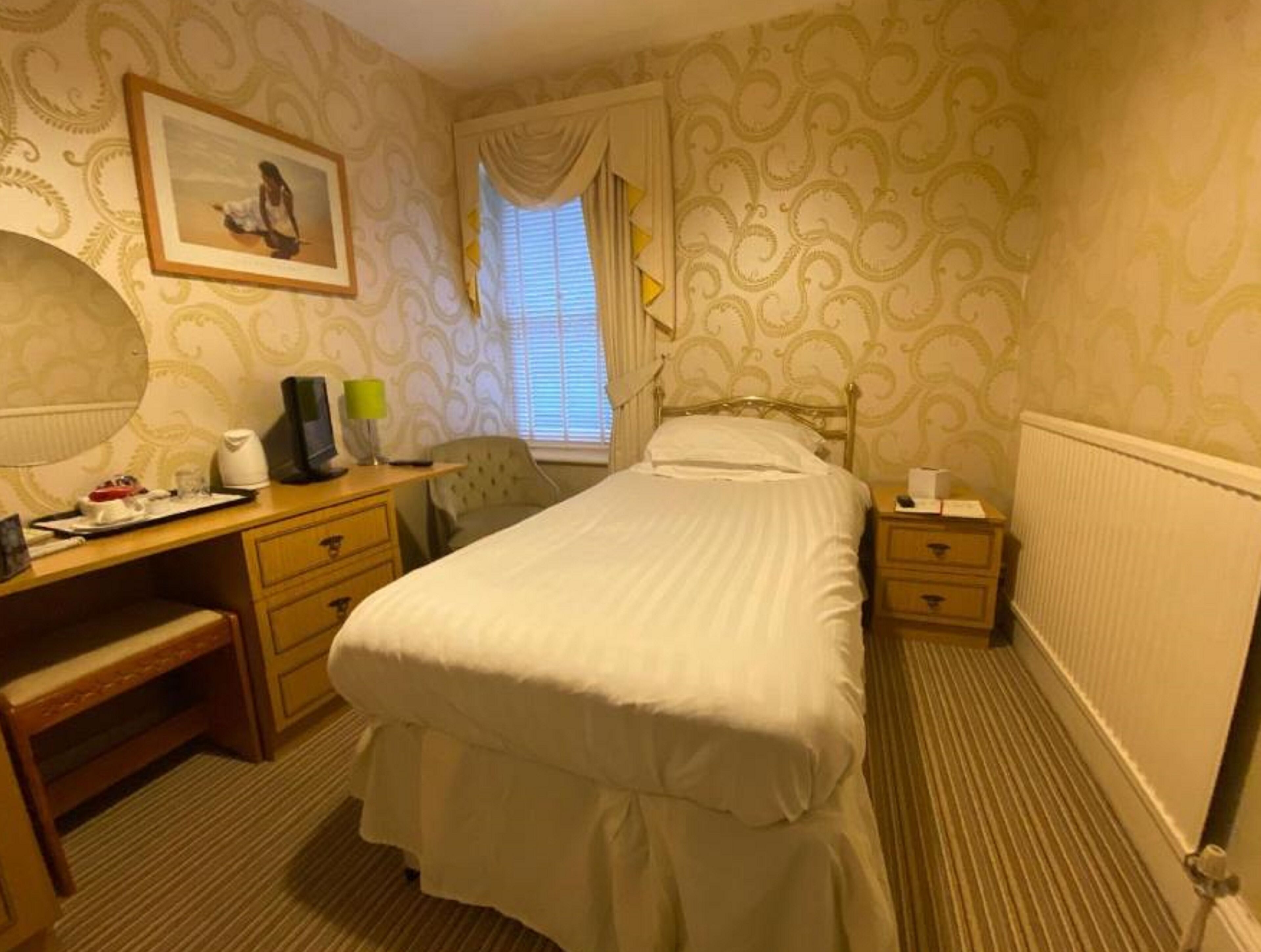 A hotel room with a bed, a desk with a chair, a bedside table, a radiator, and a window with curtains.