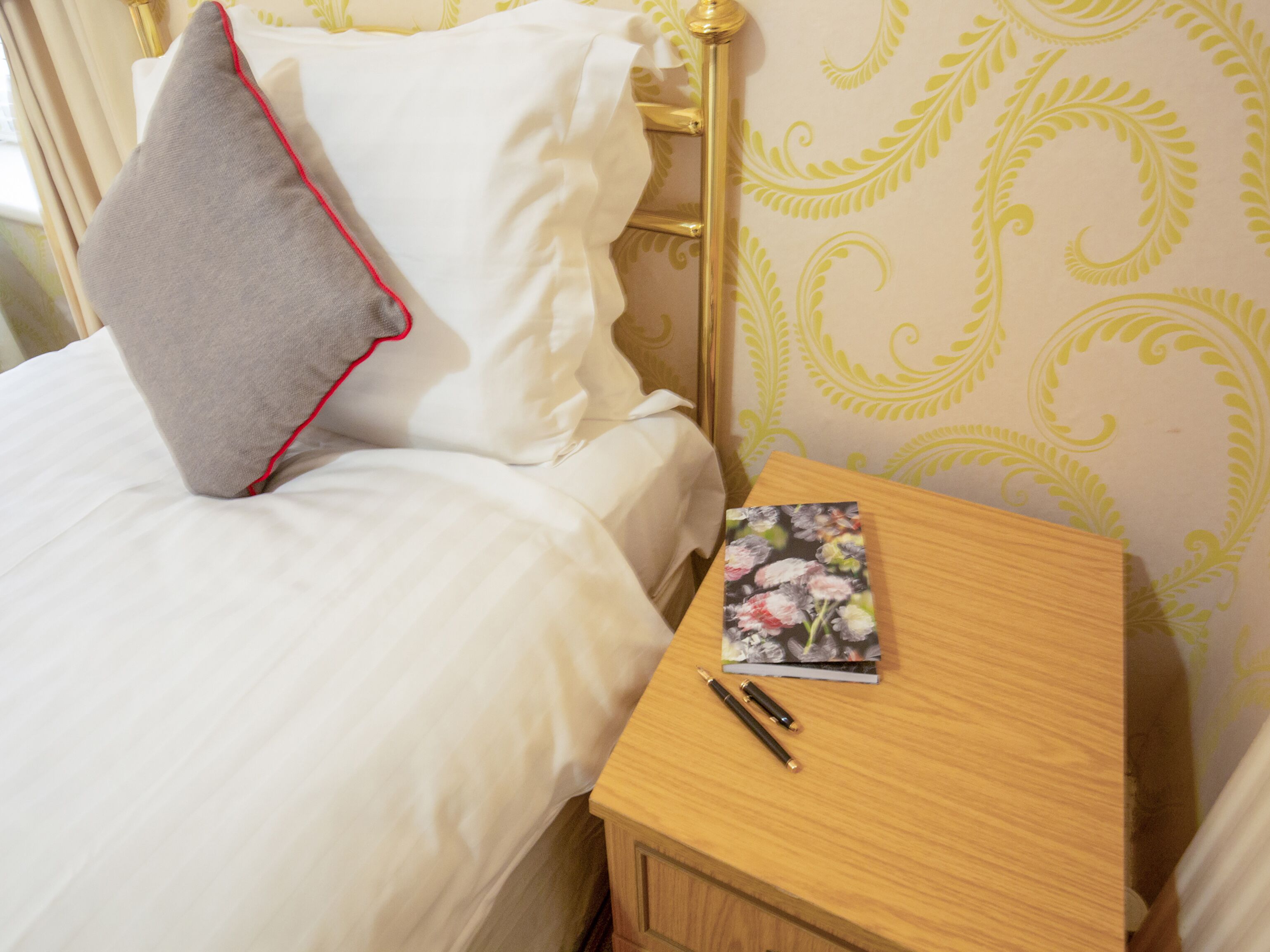 Una cama bien hecha con sábanas blancas y un almohadón gris con detalles rojos. Una mesita de noche de madera con un libro floral y dos bolígrafos se encuentra al lado de la cama.