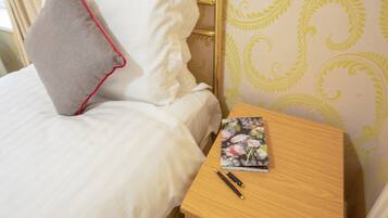 A neatly made bed with white linens and a grey pillow with a red trim. A wooden nightstand with a floral book and two pens is beside the bed.