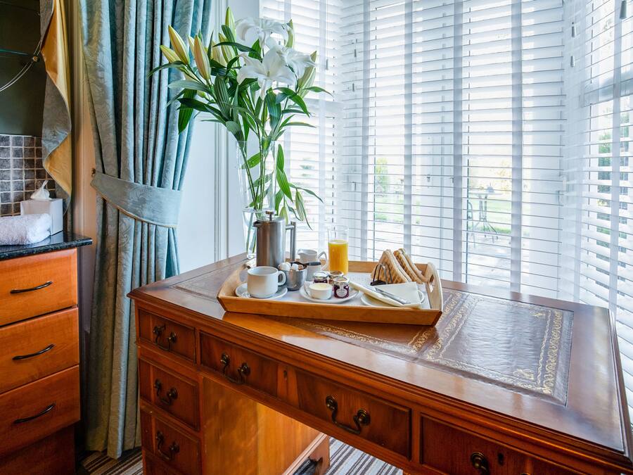A wooden desk with a tray of breakfast items, a vase with white flowers, and a window with blinds.