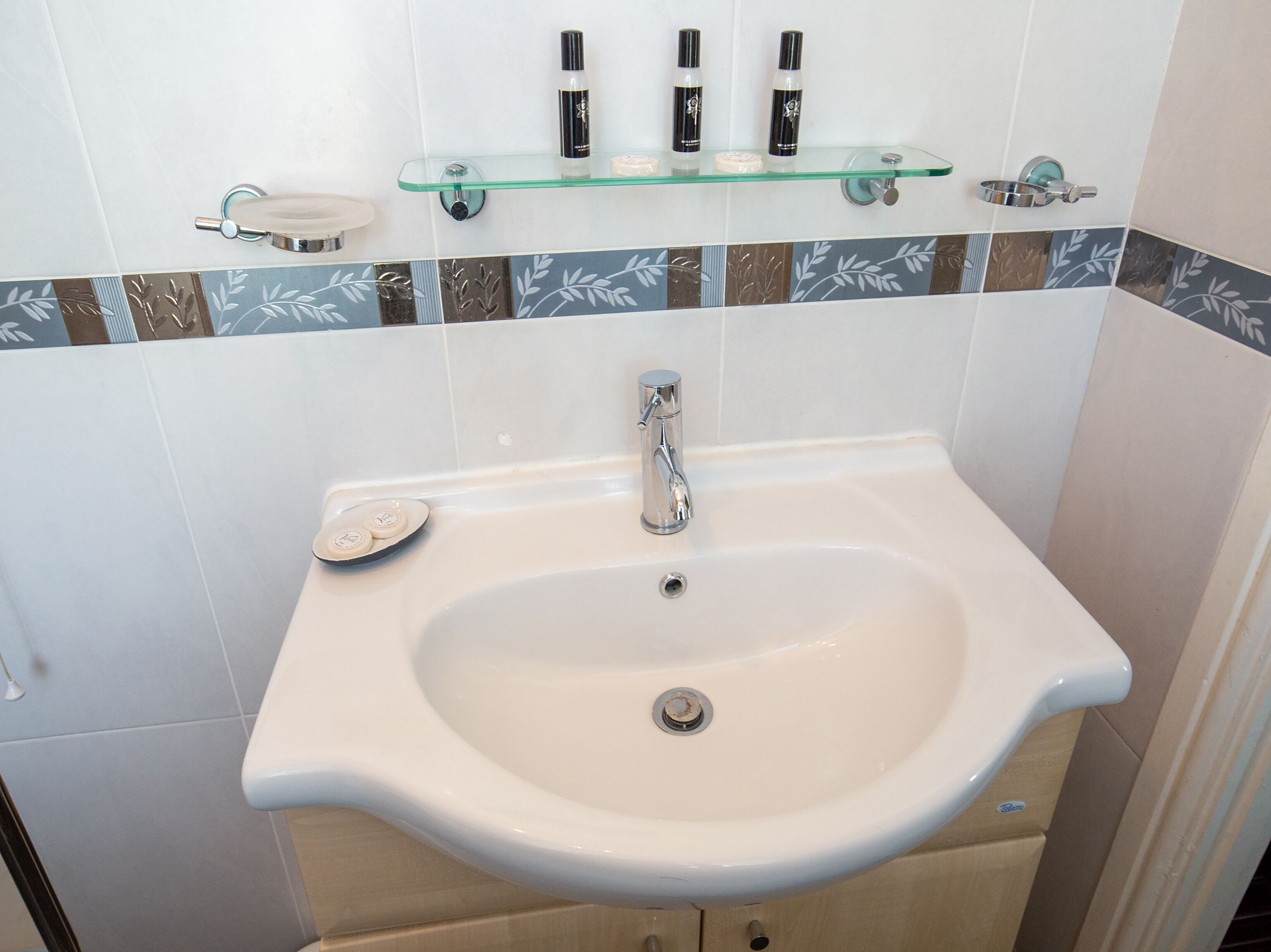 Un baño con un lavamanos blanco, un estante de vidrio encima y un salpicadero de azulejos con diseño de hojas.