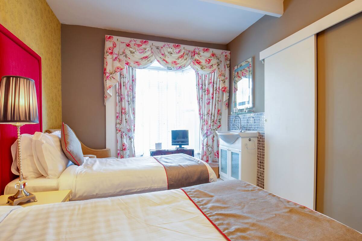 A hotel room with a bed, a desk, a lamp, a TV, and a bathroom with a sink and mirror.