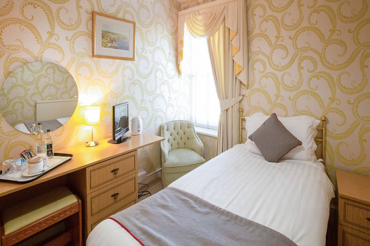 A hotel room with a bed, bedside table, desk, chair, and a window with curtains.