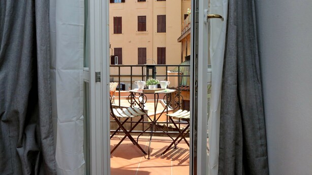 Ein Balkon mit Tisch und Stühlen, einem kleinen Topfpflanzen und Blick auf ein Gebäude mit vielen Fenstern.