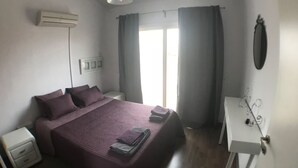 A bedroom with a bed, a nightstand, a bedside table, an air conditioning unit, a window with curtains, and a wall-mounted mirror. - Unique 2 bedroom modern apartment (Ayia Napa)