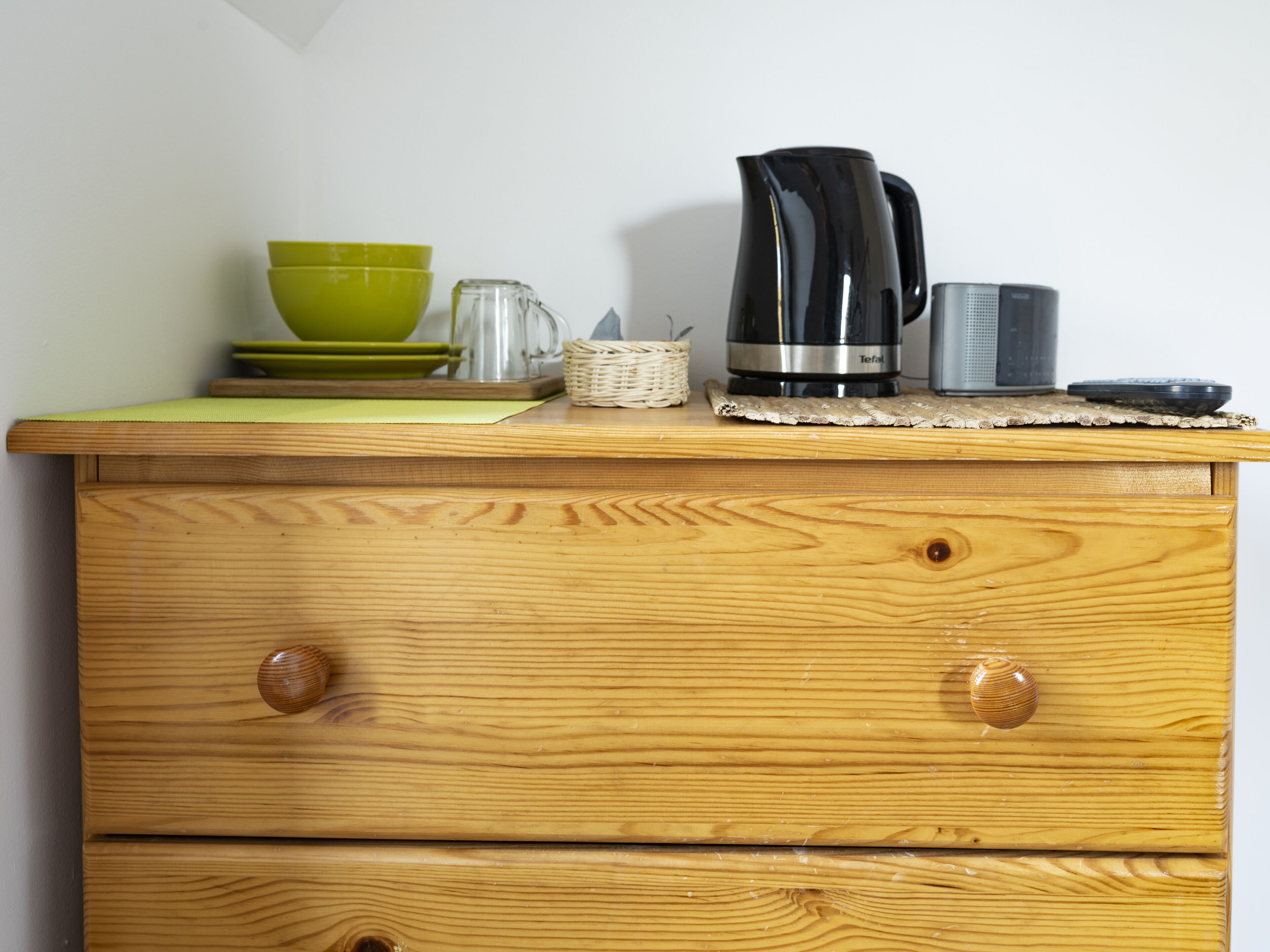 Un aparador de madera con dos cajones y una superficie con utensilios de cocina, incluyendo un bol verde, una jarra de vidrio, una cafetera negra y un recipiente gris.