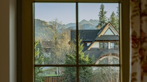 A window with a view of a house with a sloped roof, surrounded by trees and mountains. - Willa Ślimakówka (Zakopane)