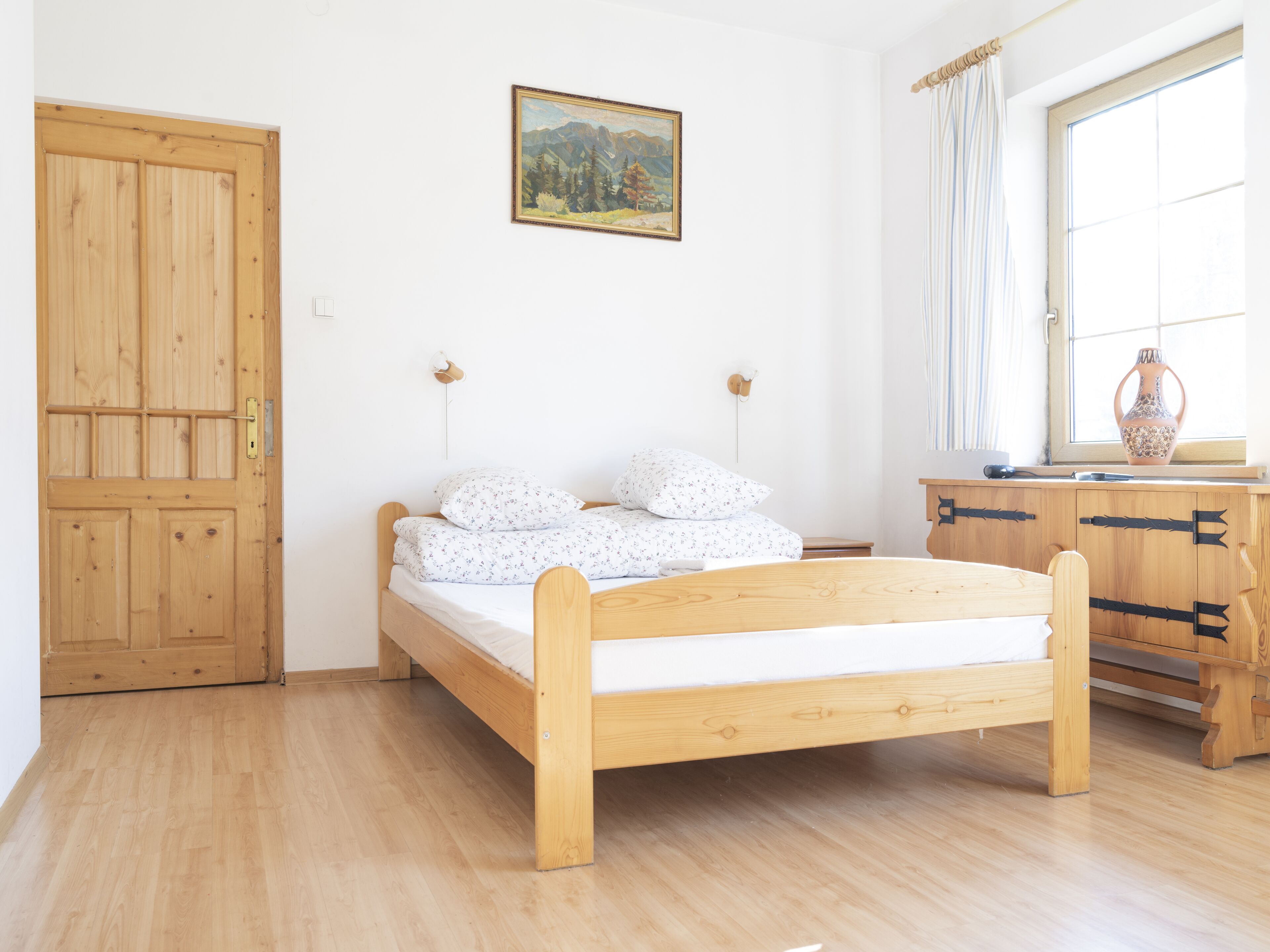 Um quarto com cama de madeira, cómoda de madeira, um quadro na parede e janela com cortinas.