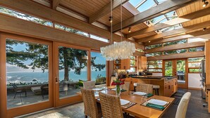 A modern kitchen with wooden cabinets and a large island, a dining area with wicker chairs and a wooden table, and a view of the ocean through large glass doors. - Sanctuary House - Architectural Gem w/ Ocean Views! (Trinidad)