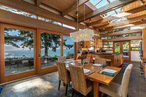 A modern kitchen with wooden cabinets and a large island, a dining area with wicker chairs and a wooden table, and a view of the ocean through large glass doors. - Sanctuary House - Architectural Gem w/ Ocean Views! (Trinidad)
