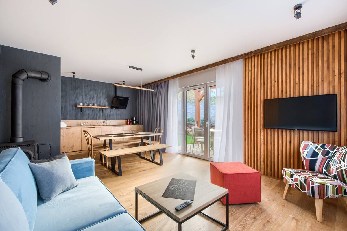 Een moderne woonkamer met een bank, salontafel en een kleurrijke stoel. Achter een glazen deur is een keuken met een eettafel en stoelen te zien.