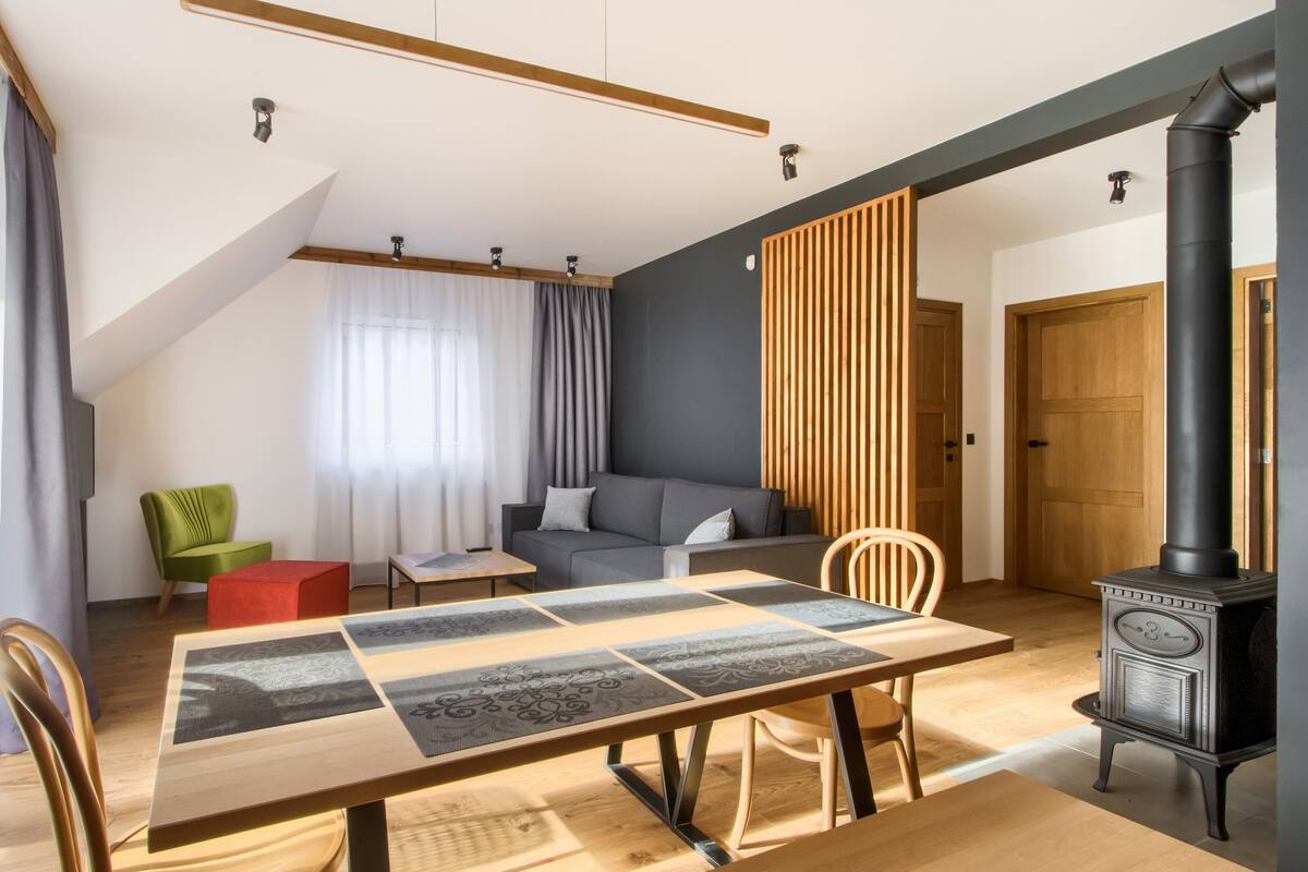 Een moderne woonkamer met een houten eettafel, een bank en een zwart houtkacheltje.