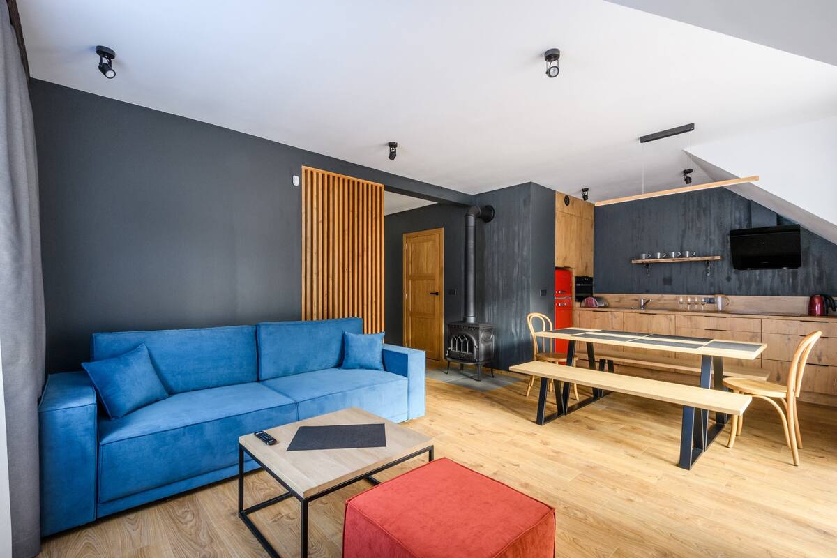 Een moderne woonkamer met een blauwe bank, een houten salontafel en een rode poef. Achtergrond toont een keuken met een houten bankje en een televisie.