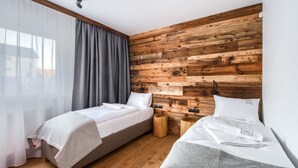 Une chambre d’hôtel moderne avec deux lits simples, des murs recouverts de panneaux en bois, une grande fenêtre avec des rideaux légers et une table de chevet avec une lampe.