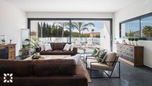 Una sala de estar moderna con un amplio ventanal corredero que da a un balcón, amueblada con un sofá de cuero marrón, un sillón color beige y una mesa de centro de vidrio.