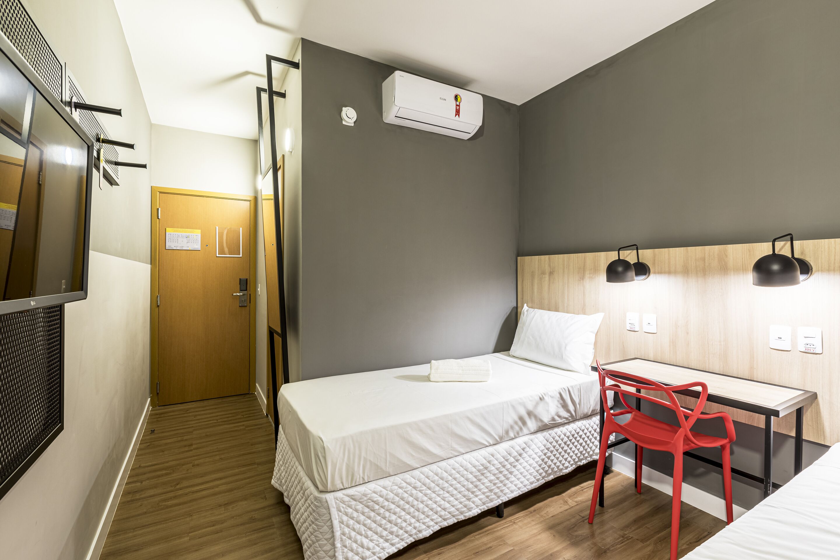 A hotel room with a bed, a desk, a chair, an air conditioning unit, and a door.