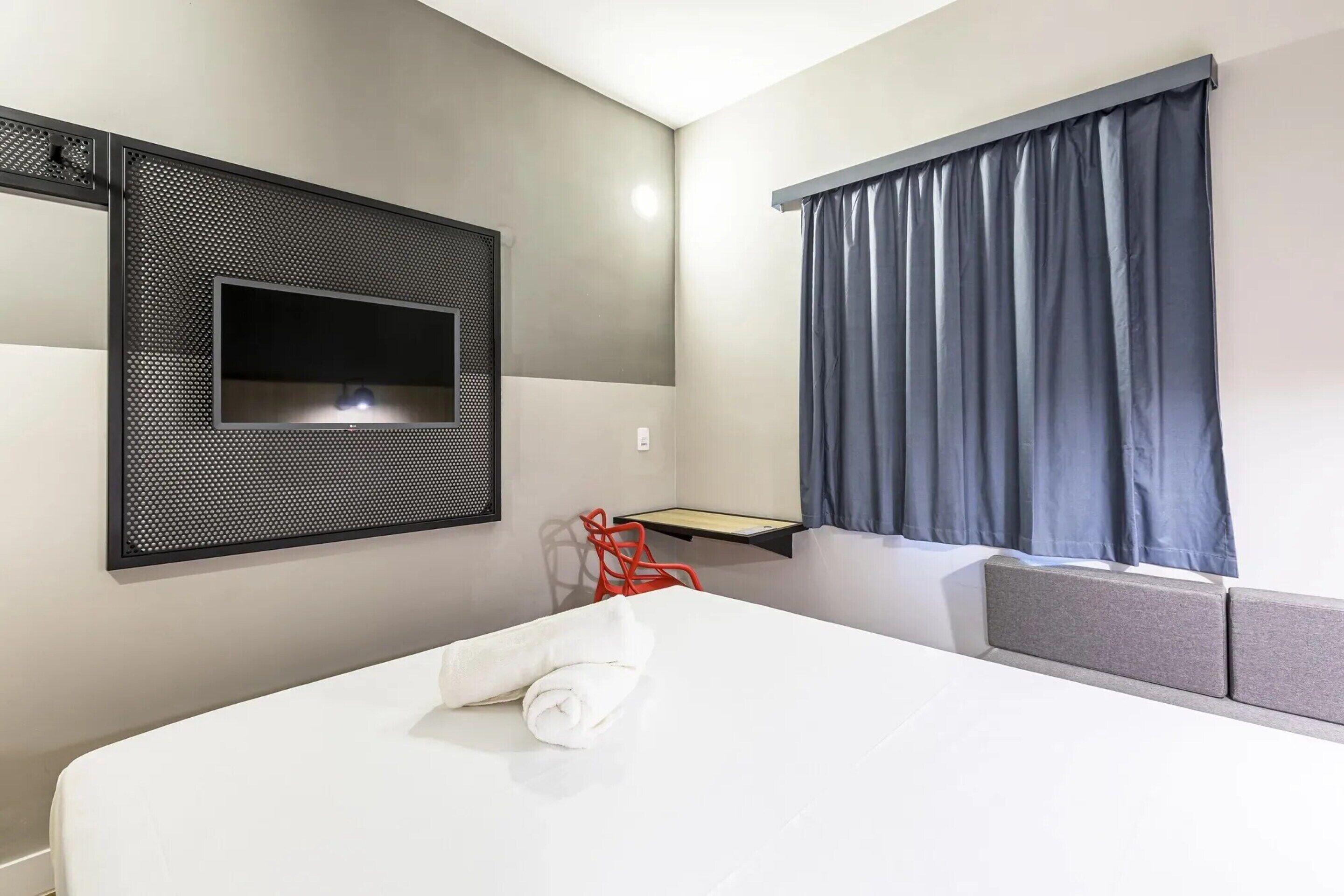 Ein Hotelzimmer mit Bett, Wandfernseher, Schreibtisch und einem fenster mit Vorhängen.