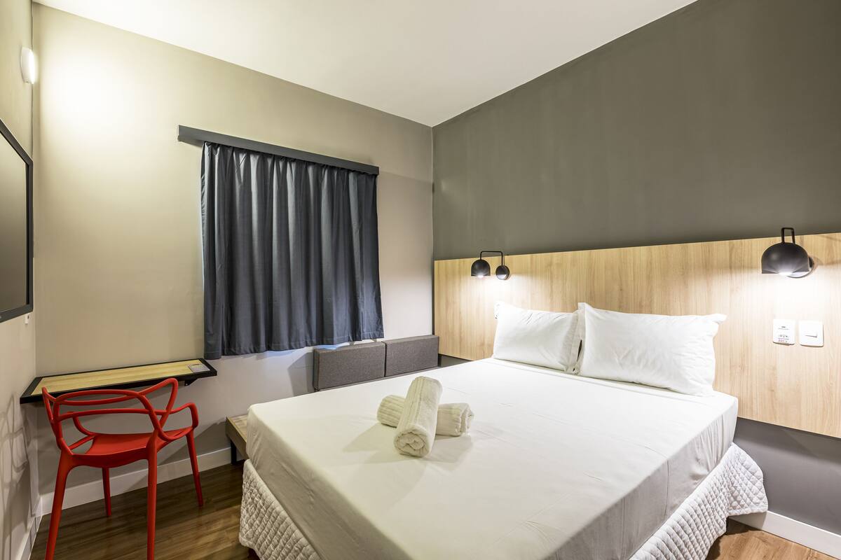 Ein Hotelzimmer mit Bett, Schreibtisch mit Stuhl, Fenster mit Vorhängen und einem Kopfteil mit Wandleuchten.