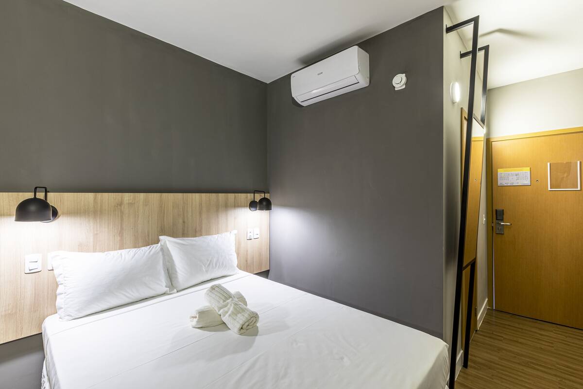 Ein Hotelzimmer mit einem Bett, weißen Leinentextilien, einem Holzkopfteil, einer Klimaanlage, einer Wandleuchte und einer Holztür.