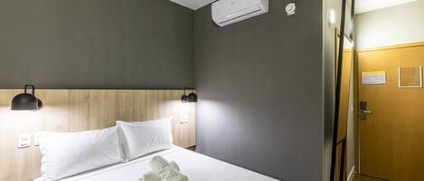 Habitación de hotel con cama, ropa blanca, cabecera de madera, unidad de aire acondicionado, lámpara de pared y puerta de madera.