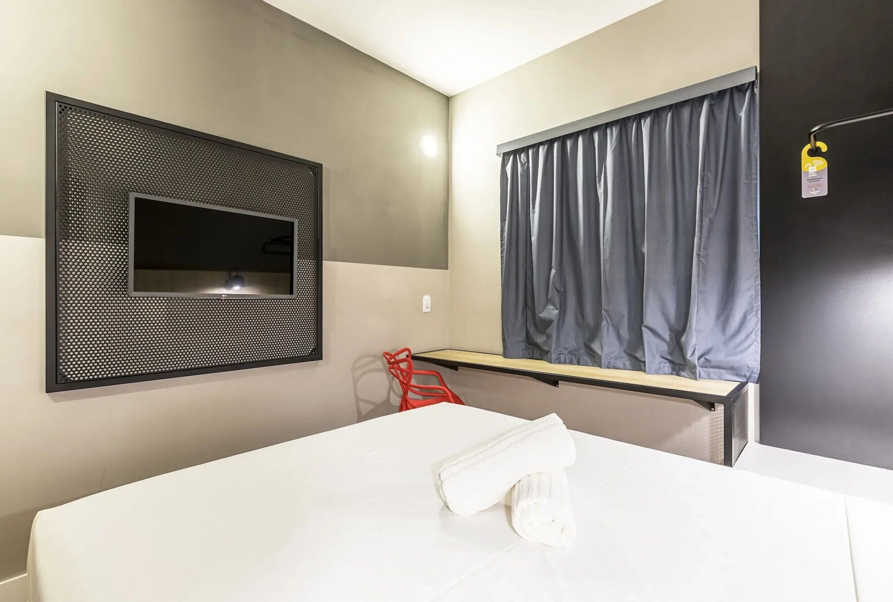 Una habitación de hotel con cama, televisión montada en la pared, ventana con cortinas y una pequeña mesa.