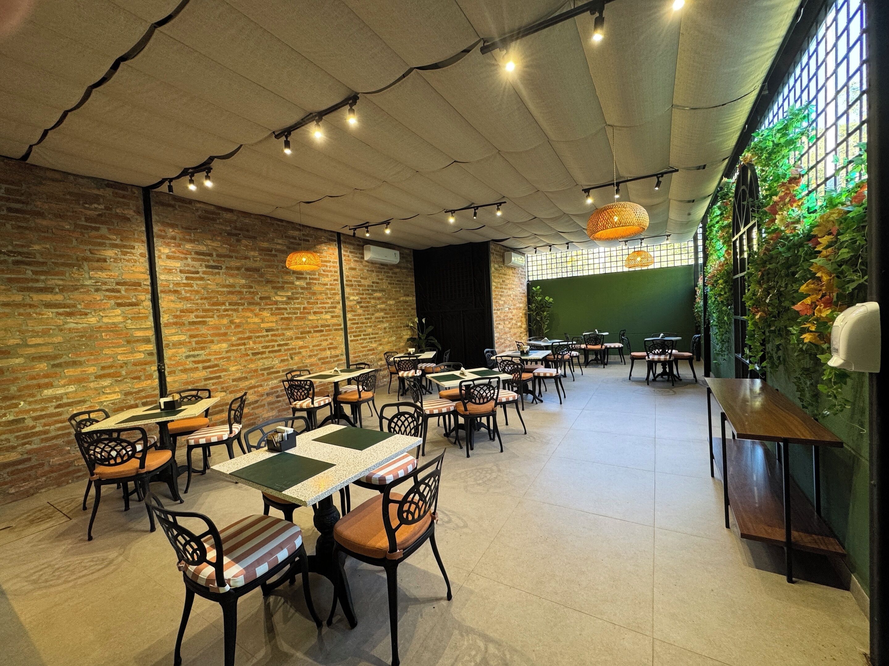 A restaurant with brick walls, wooden tables, and chairs. There are hanging lights and a green wall with plants.