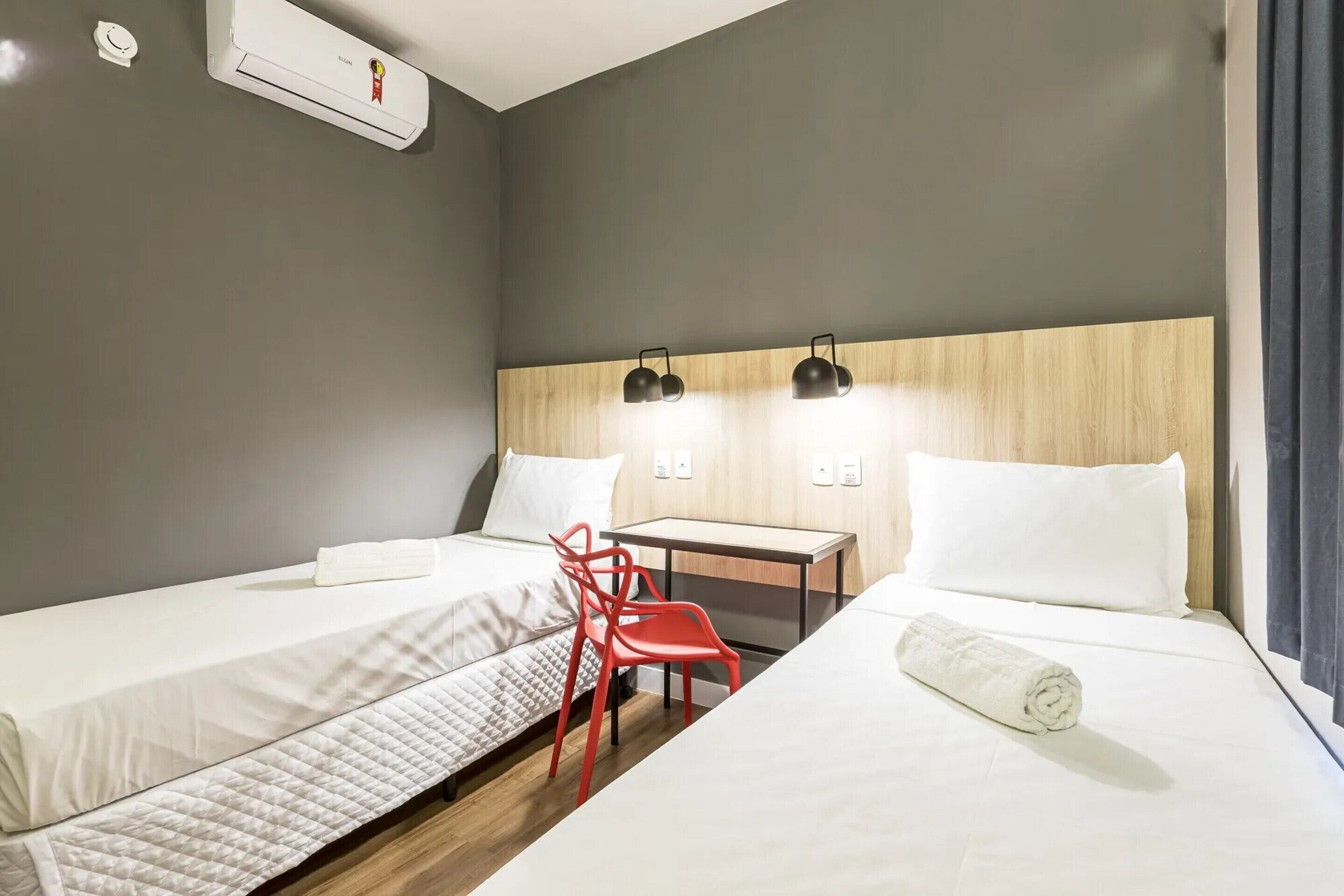 Habitación de hotel con dos camas, un escritorio, una silla roja y aire acondicionado instalado en la pared.