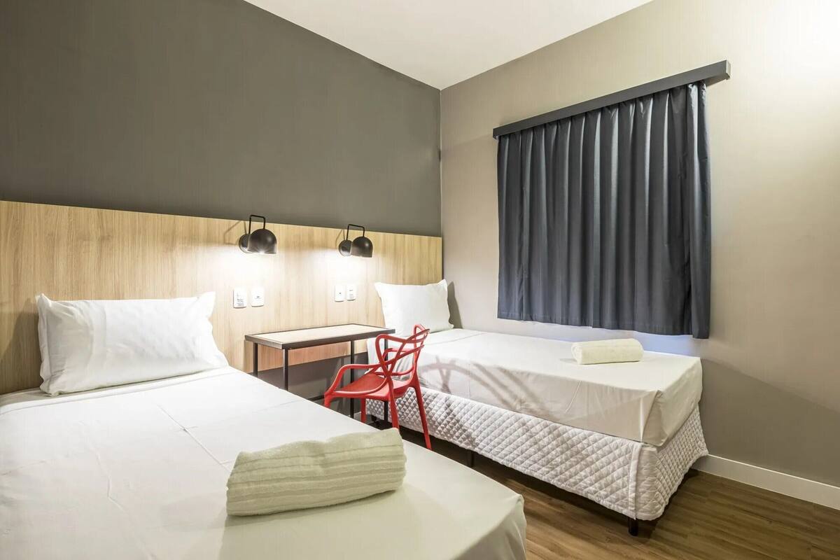 Ein Hotelzimmer mit zwei Betten, einem Schreibtisch, einem Stuhl und einem Fenster mit Vorhängen.