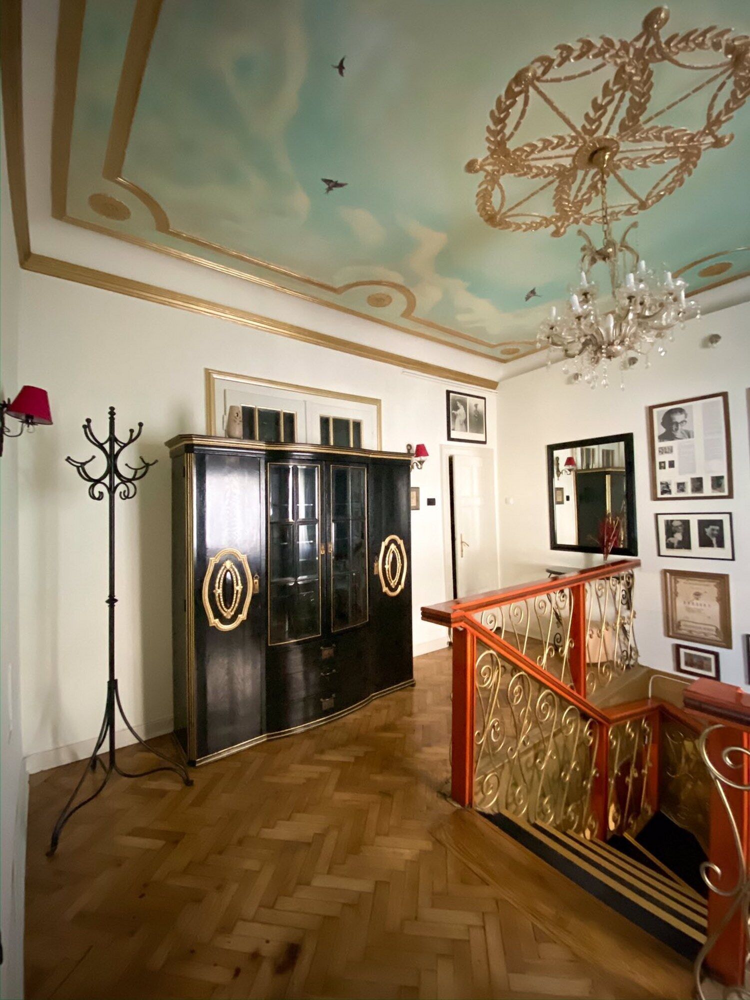 A room with a wooden herringbone floor, a black wardrobe with gold accents, a red and gold railing, and a chandelier.