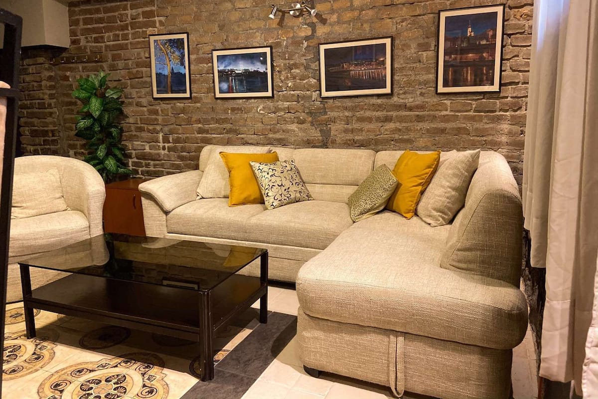 Una sala de estar con pared de ladrillo, un sofá color beige con cojines amarillos, una mesa de centro de vidrio y cuadros enmarcados en la pared.