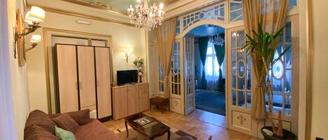 A spacious living room with a sofa, a coffee table, and a TV. There is a chandelier, a potted plant, and a view of the outside through a glass door.