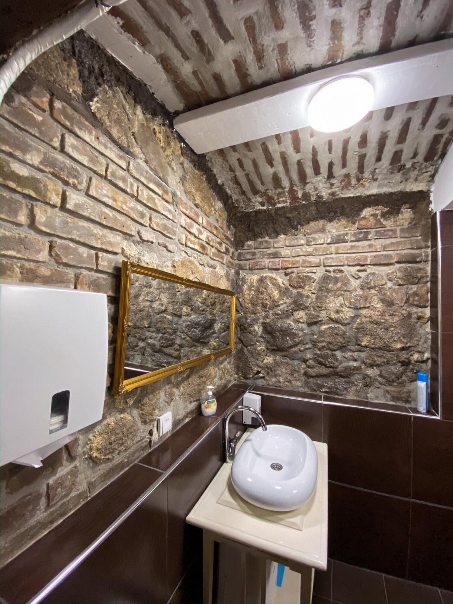 Une salle de bain avec un mur en pierre, un miroir avec un cadre doré, un lavabo blanc et un sèche-mains.