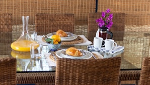 A breakfast setting with wicker chairs, a glass table, plates with croissants, a bowl of fruit, a vase with flowers, and a coffee pot. - Villa Near the Beach (Paralimni)