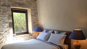 Una cama con ropa de cama blanca y almohadas a rayas, dos lámparas de noche, una ventana con vistas a zonas verdes y una pared de piedra.