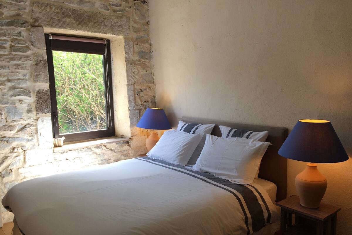 A bed with white bedding and striped pillows, two bedside lamps, a window with a view of greenery, and a stone wall.