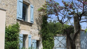 Un edificio de piedra con contraventanas azules, un sendero de piedra y un árbol con flores rosadas.