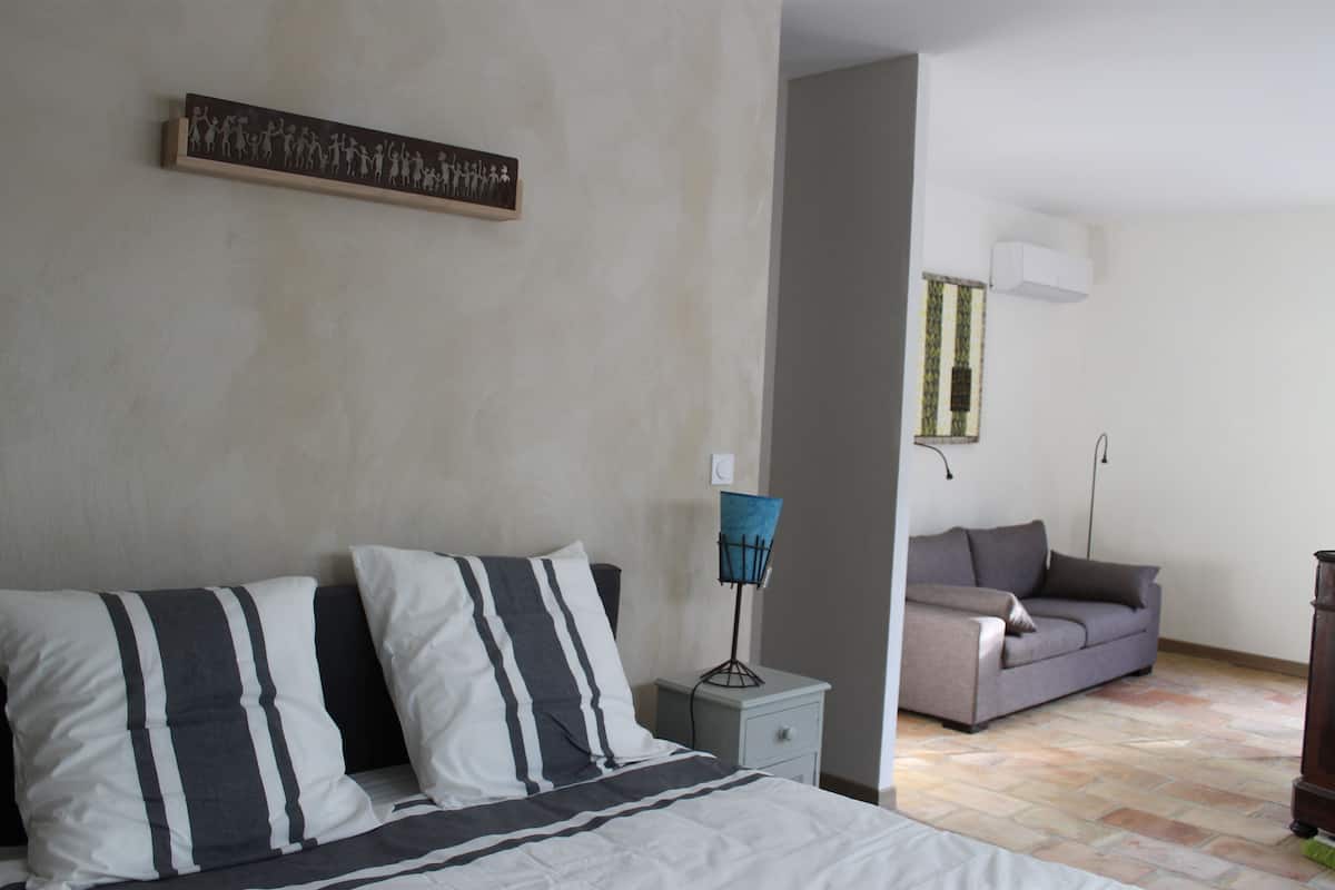 A bedroom with a bed, a nightstand, a lamp, a sofa, and a wall-mounted artwork.