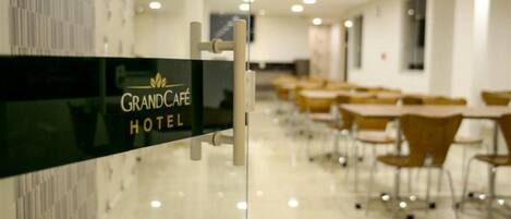 A Grand Café Hotel with a modern interior featuring a long row of tables and chairs, a glass partition with the hotel's name, and a clean, well-lit environment.