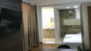 Habitación de hotel moderna con televisión de pantalla plana, una mesa de comedor, una silla de mimbre, una pequeña planta en maceta y un baño con mampara de vidrio y una cama.