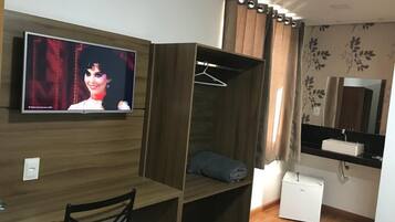Una habitación con un escritorio de madera, un televisor montado en la pared, un armario, un espejo y un pequeño electrodoméstico blanco.