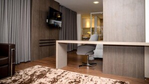 Una habitación de hotel moderna con piso de madera, un escritorio con silla, un televisor de pantalla plana en la pared y un amplio ventanal con cortinas.