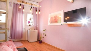 Ein Schlafzimmer mit einem Bett, einem an der Wand befestigten Fernseher, einem Fenster mit Vorhängen und einer Zimmerpflanze.